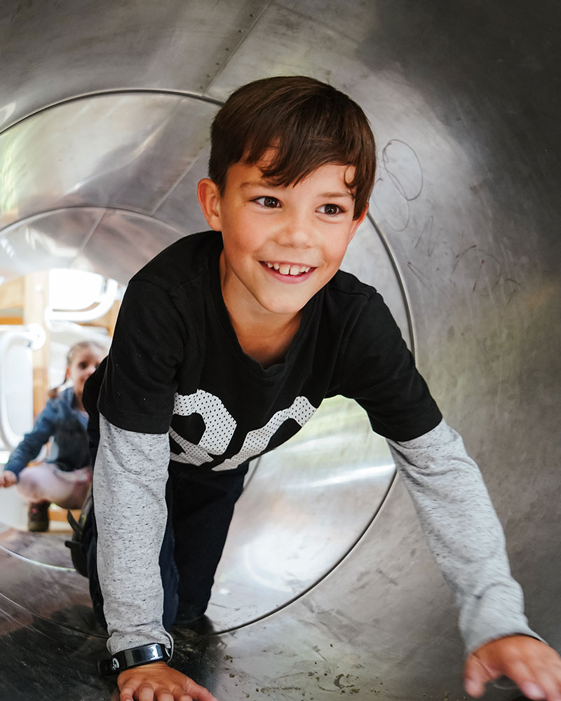 A young boy is climbing through a steel playground crawl tube and smiling.